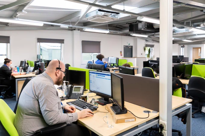 Modern office space with employees working at desks equipped with dual monitors and green office chairs.
