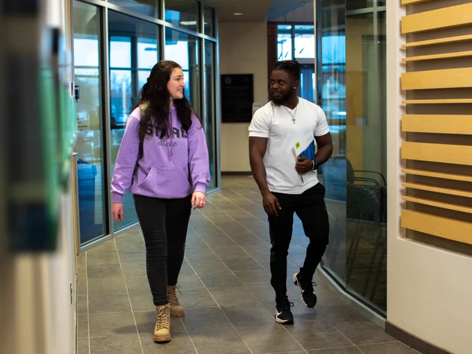 Two individuals, one wearing a purple "Stark State" hoodie, walking and talking in a brightly lit indoor hallway.