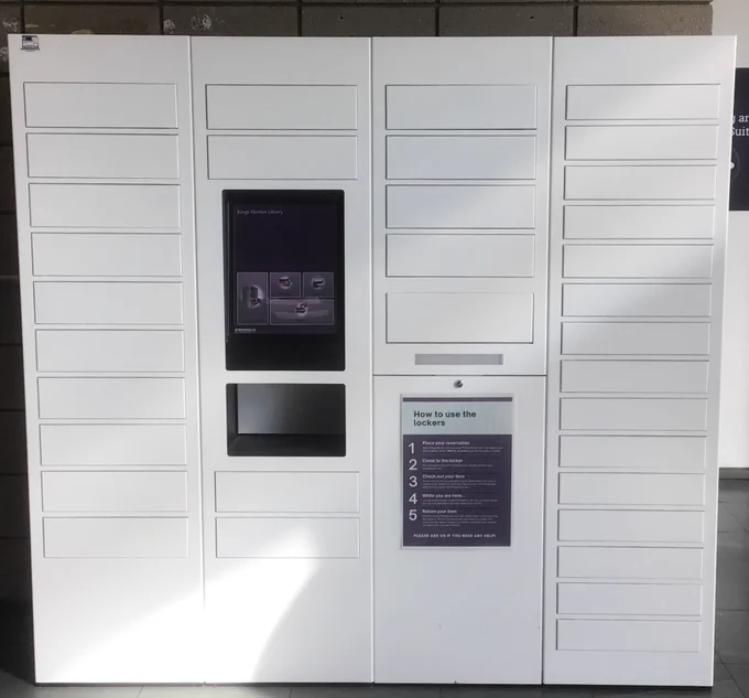 White automated locker system with multiple compartments and a touchscreen interface labeled Kings Norton Library for item pickup and return.