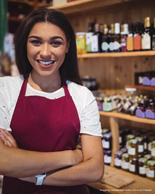 Person in a burgundy apron standing in front of shelves stocked with various products, smiling at the camera.