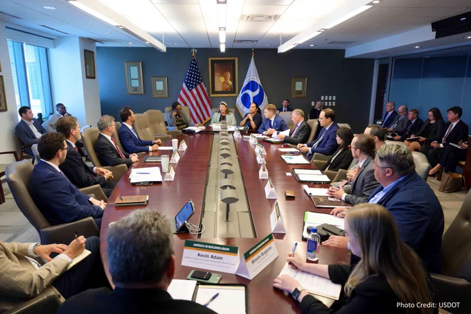 Photo of a formal meeting at a long table with multiple participants, featuring the flags of the United States and the Department of Transportation prominently displayed. Credit: USDOT
