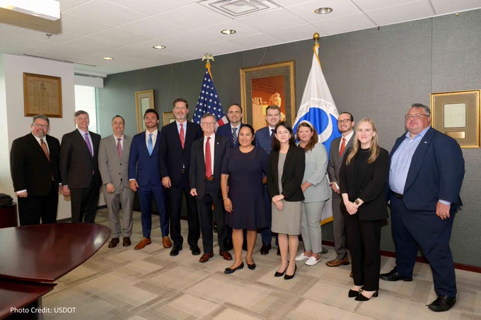 Group of fourteen individuals posing for a photo in a conference room with flags of the United States and the Department of Transportation, along with framed images on the wall.
