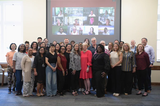 Photo with several dozen people posing with Maureen Conroy, Ph.D., and Patricia Snyder, Ph.D., at their UF College of Education retirement party. Behind the group, a screen shows additional people on Zoom. There is a lot of light coming in from behind the group due to the placement of windows in the room. 
