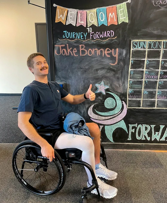 Jake Bonney gives a thumbs-up while seated in a wheelchair at Journey Forward, a neuro-recovery center. Behind him is a colorful chalkboard welcoming him by name.
