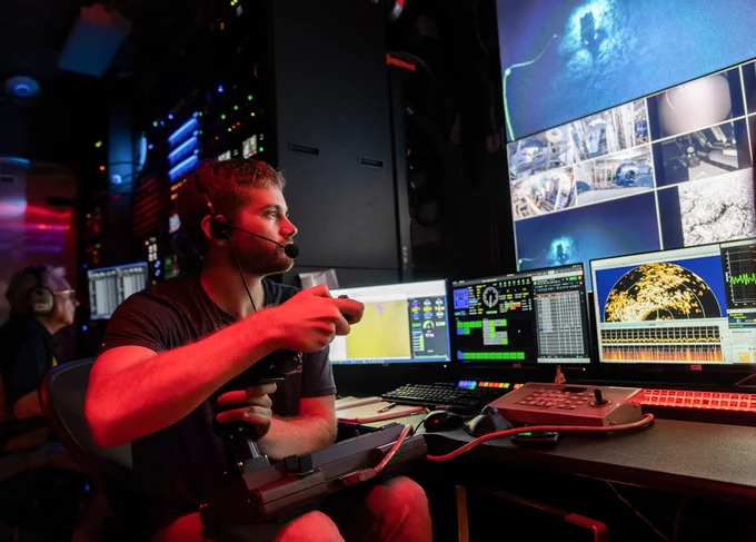 Jake Bonney sits at a console in the Ocean Exploration Trust control van, wearing a headset and operating a joystick. Multiple monitors in front of him display data and underwater imagery from an ROV.