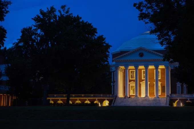 Night view of the Rotunda at the University of Virginia, illuminated by warm lights against a twilight sky.