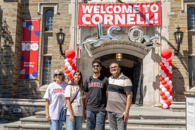 Four individuals pose happily in front of a decorated entrance at Cornell University, with a "Welcome to Cornell" banner and red and white balloons.