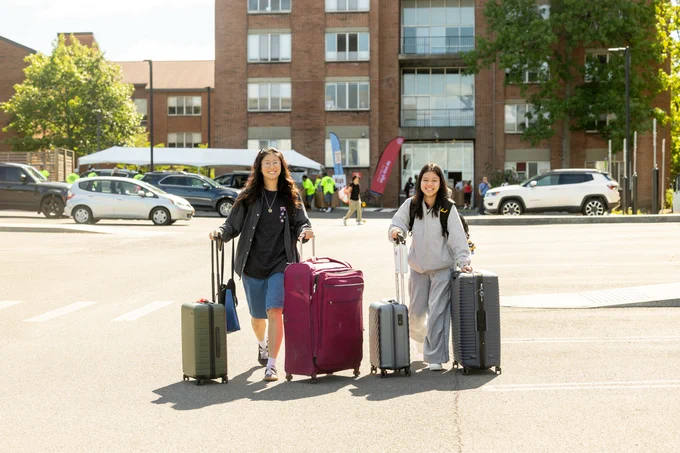Two individuals each holding luggage, smiling and standing in a sunny parking lot, with buildings and parked cars in the background.