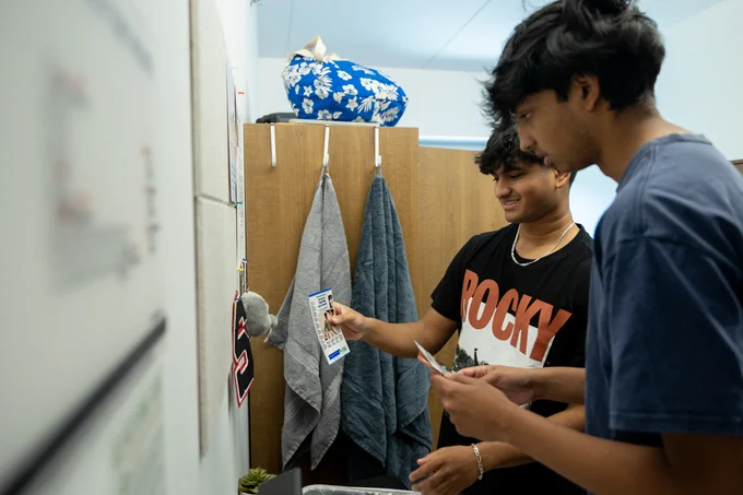 Two people collaborating on a project in a dorm room, one holding a textbook and the other writing notes, with personal items hanging on a wall.