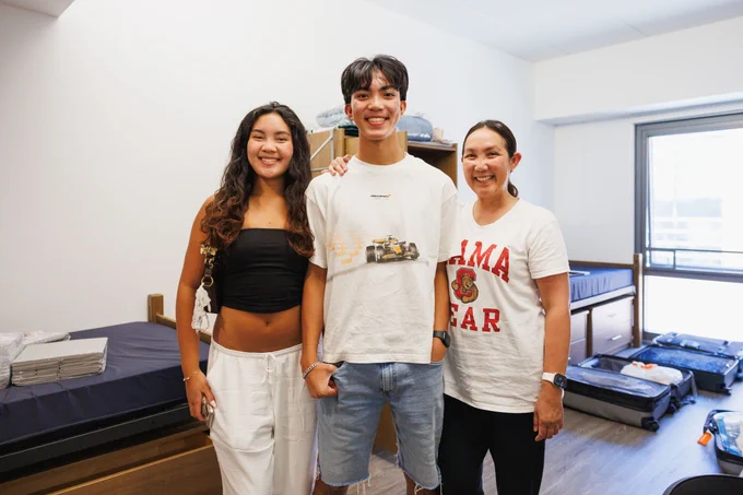 Three individuals smiling in a dorm room, with one wearing a shirt that reads "Mama Bear".