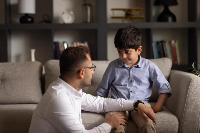 Two individuals having a conversation on a sofa in a living room, with one appearing to be an adult and the other a child. The setting includes a tastefully decorated room with a couch and various decorative items on shelves in the background.