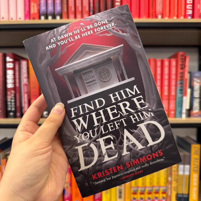 Someone holding up a copy of Find Him Where You Left Him Dead by Kristen Simmons in front of bookshelves filled with red and yellow books.