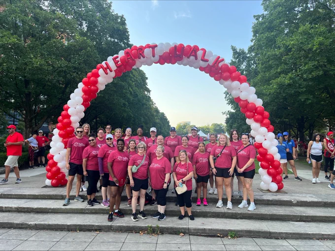 A group of Heart Walk Quantum Health Healthcare Warriors® in matching red shirts pose together under a red-and-white balloon arch spelling out “HEART WALK,” with trees framing the background.