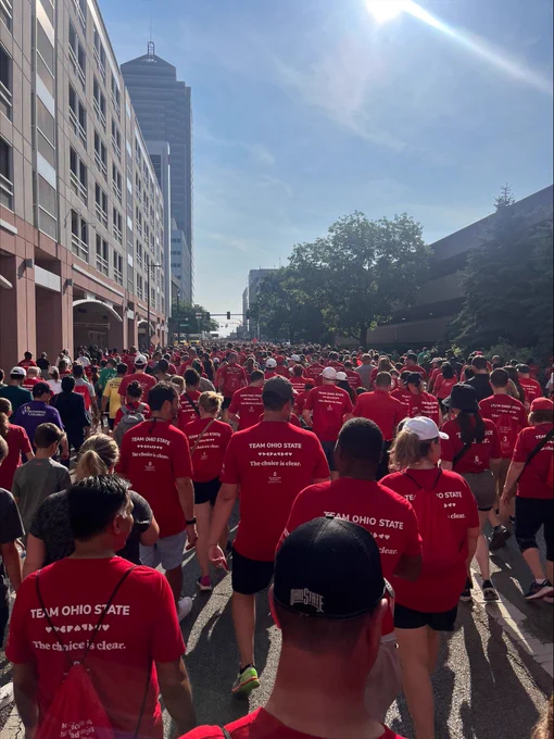 A wide crowd of participants intermingled with Quantum Health Healthcare Warriors® in matching red “Team Ohio State” shirts walk down a sunlit city street during the Heart Walk, with tall buildings and bright morning sunlight overhead.