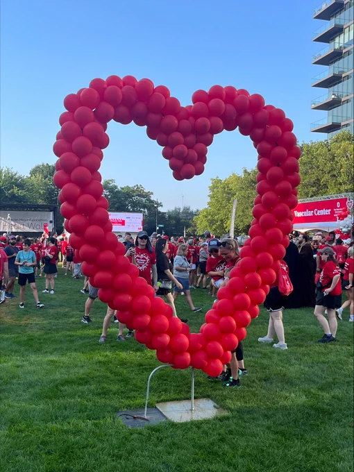 A large red heart made of balloons stands on a grassy field at the 2025 American Heart Association Heart Walk event, with participants in red shirts gathering in the background under a clear blue sky.