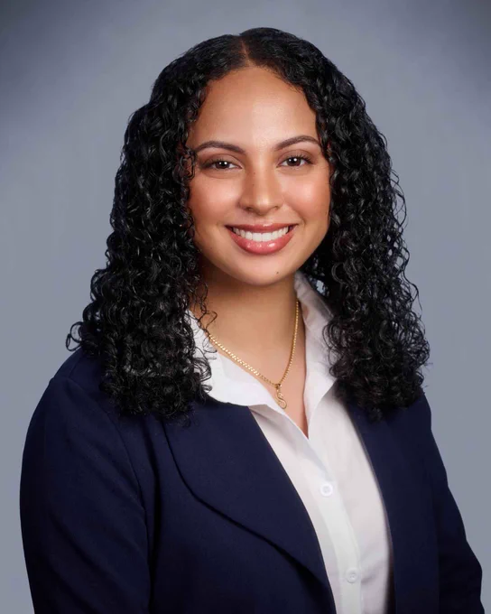 Professional headshot of a person with curly hair, wearing a navy blazer and white shirt, smiling against a grey background.