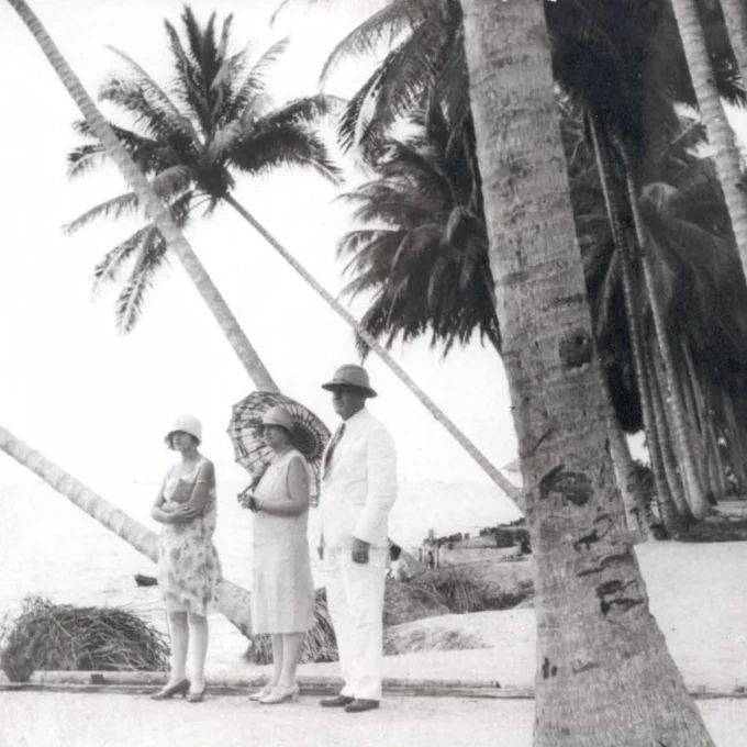 Three individuals standing under palm trees on a beach, with one holding an umbrella.