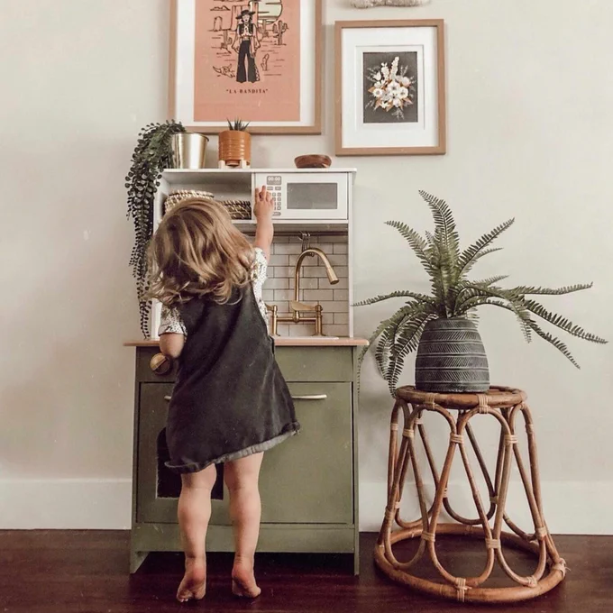 A child plays with a toy kitchen in a room decorated with plants and vintage-style art.