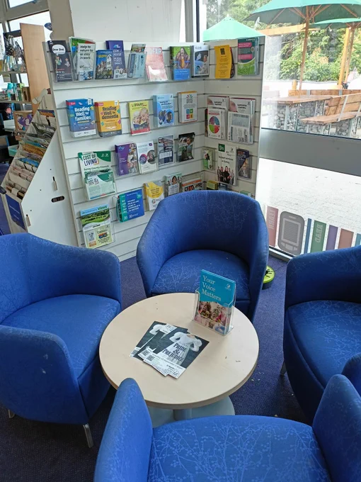 Photograph of four blue tub chairs around a small circular coffee table with a corner stand of leaflets behind them. 