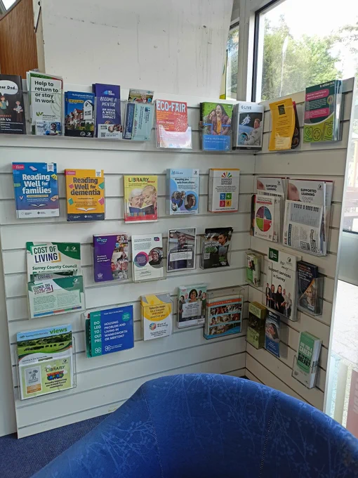 Close up photograph of a corner stand with leaflets displayed in plastic holders. There is a corner of a blue tub chair in the close foreground. 