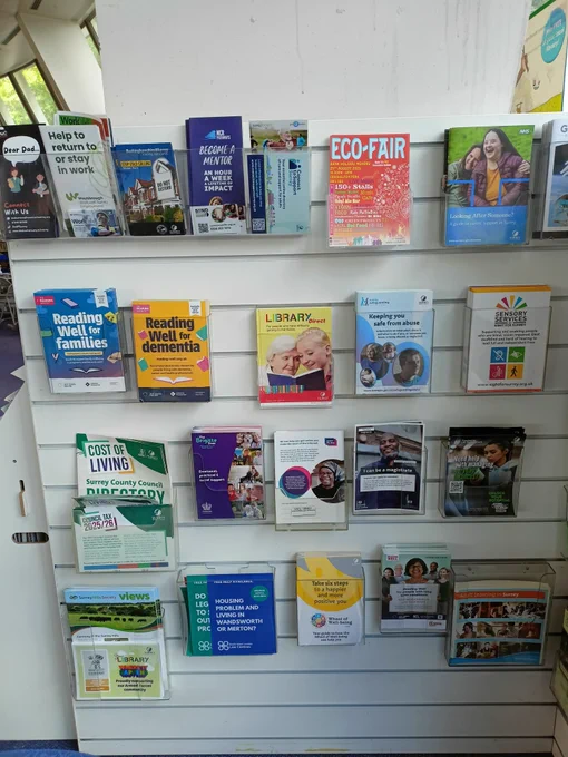 Photograph of a stand displaying information leaflets in plastic holders. The leaflets include information on Cost of Living, Reading Well for Families and Reading Well for Dementia, and A Guide to Carers' Support in Surrey.