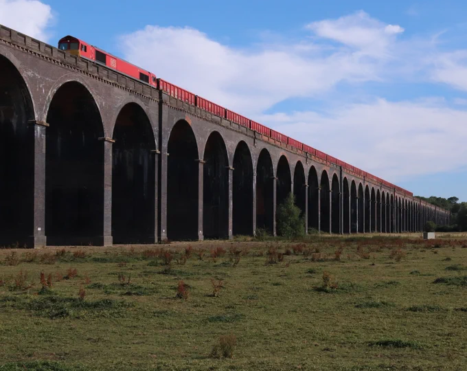 A red train passing over a Welland Viaduct, a long, multi-arched stone viaduct in a rural landscape under a partly cloudy sky.