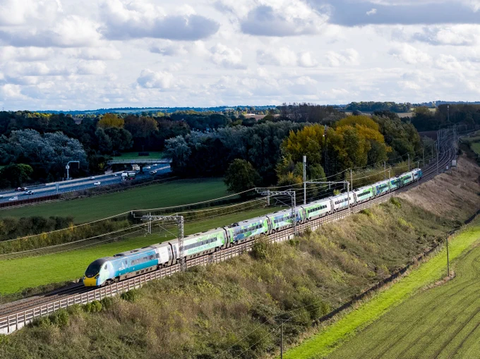 An Avanti West Coast train traveling through a countryside landscape with lush green fields and scattered trees under a partly cloudy sky.