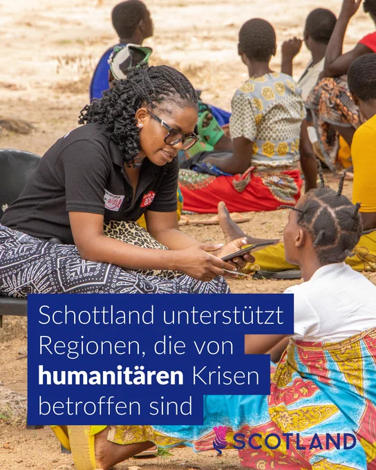 „Eine Gruppe von Menschen sitzt im Freien auf dem Boden. Im Vordergrund interagieren zwei Personen miteinander – eine mit geflochtenen Haaren und Smartphone, die andere in bunter Kleidung. Im Hintergrund sind weitere Personen zu sehen. Am unteren Bildrand steht der Text: ‚Schottland unterstützt Regionen, die von humanitären Krisen betroffen sind.‘ Neben dem Text befindet sich ein Logo mit der Aufschrift ‚SCOTLAND‘.“