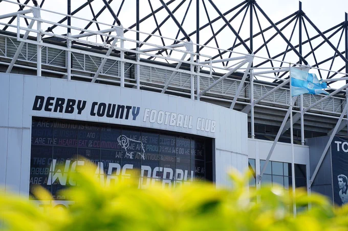 Pride Park Stadium entrance with "Derby County Football Club" sign, crisscross stadium roof structure, and a fluttering Derbyshire flag. Foreground shows blurred green leaves.