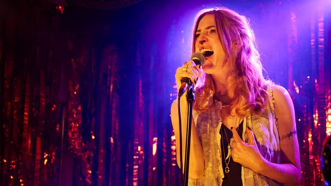 A woman with long red hair stands on stage, singing into a microphone