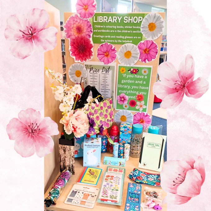 Photo of the Library Shop display at Ash Library, which is decorated with a summer, floral theme. The photo is on a pale pink background with images of watercolour blossoms on the border. 