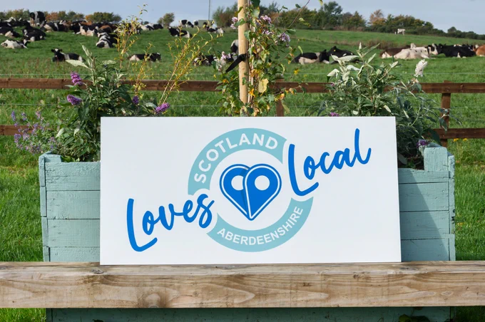 Photograph courtesy of Aberdeenshire Council.
The Scotland Loves Local Aberdeenshire banner is displayed in front of a painted tattie box of flowers, in front of a field of dairy cows at Forest Farm Dairy, Aberdeenshire.