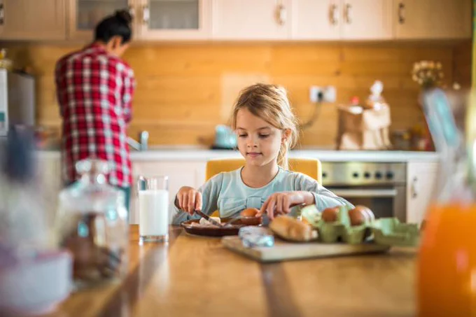 A child sits at a kitchen table, concentrating on preparing a sandwich. In the background, an adult is standing by the counter, facing away. The kitchen is warmly lit and has wooden cabinets.