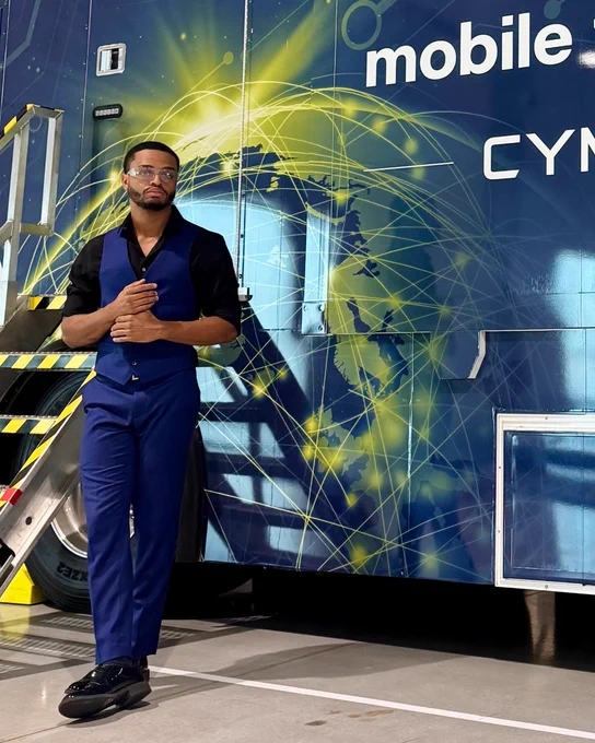 Person in a stylish blue suit standing confidently in front of a mobile threat defense, CyManII-branded vehicle, featuring a vibrant graphic of a world map in the background.