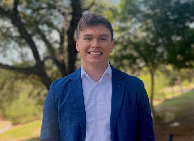 Person smiling outdoors, wearing a blue blazer, white shirt, and beige pants, standing in a park-like setting with trees in the background.