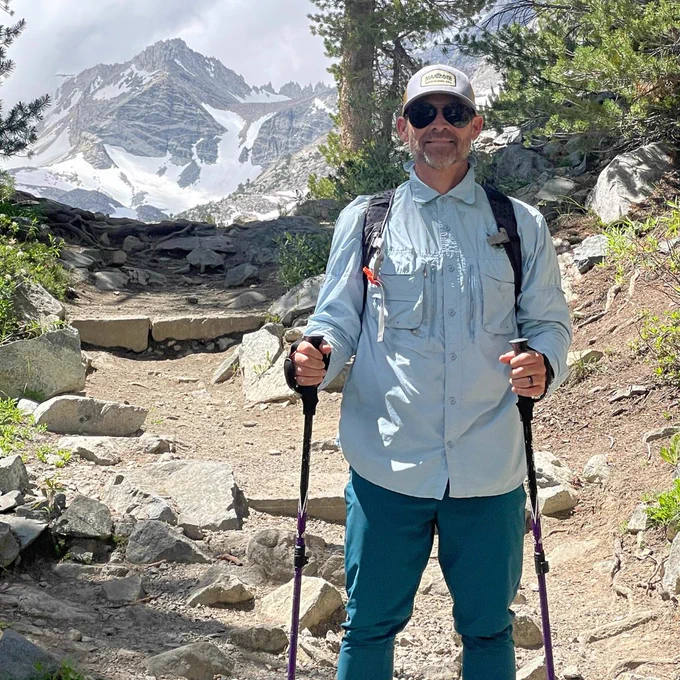 A man (Russell) wearing a light blue button-up shirt, green hiking pants, sunglasses, and a hat smiles at the camera. He is holding two hiking poles and stands on a rocky mountain path. In the background, there is a large, snow-capped mountain peak.