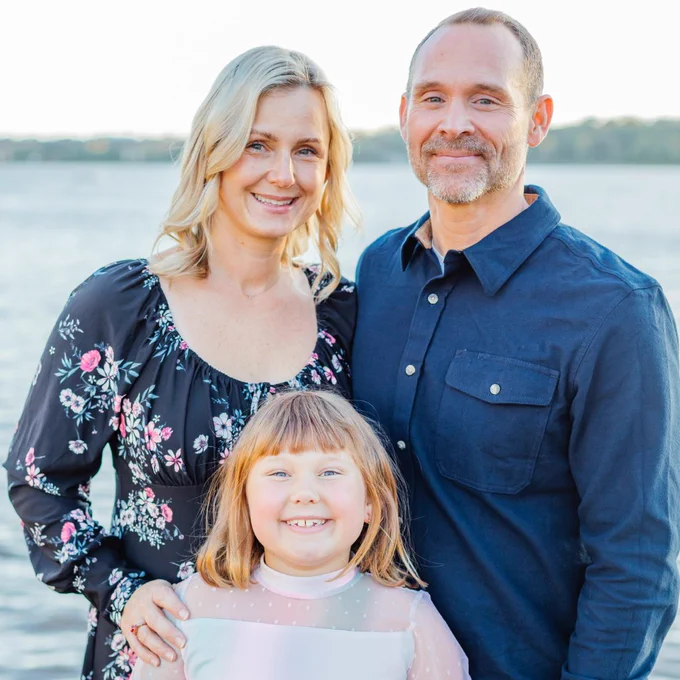 A family of three smiles at the camera for a portrait. A man in a dark blue shirt (Russell) stands on the right, with a woman in a black floral dress on the left. A young girl with reddish-blonde hair stands in front of them, smiling. They are standing in front of a body of water at sunset.