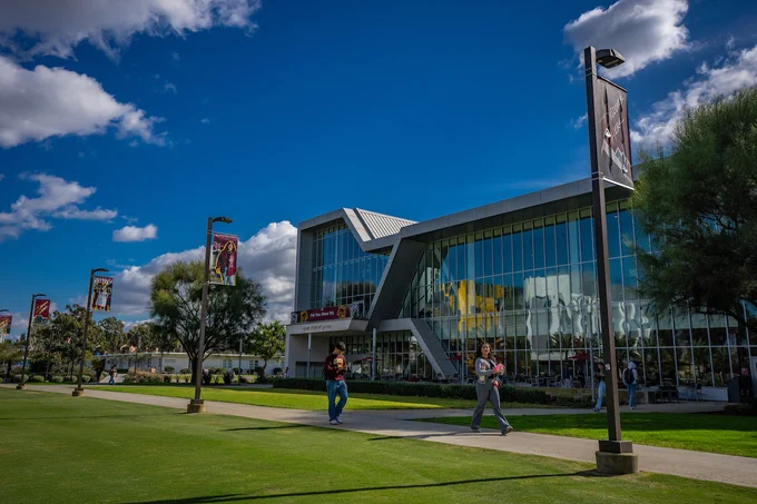 Students walk past a modern glass building with bold geometric architecture on a sunny day, banners featuring campus life hanging from lampposts along the sidewalk.