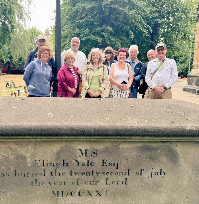 A group of a group of tour guides stood behind the tomb of Elihu Yale at St Giles Cemetery, Wrexham. A fairly bright summer's day with trees in the background 