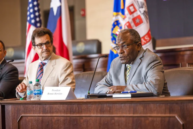 President Hawkins speaking at the microphone while seated and other members of the signing party look on. 