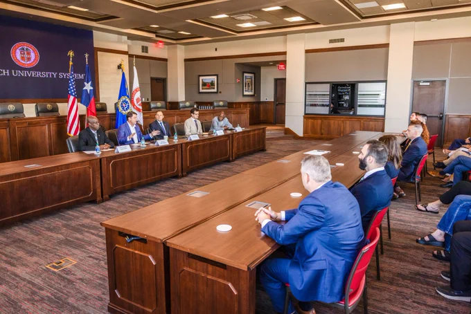 Five individuals sitting at a large table at the front of the room as audience members look on. 
