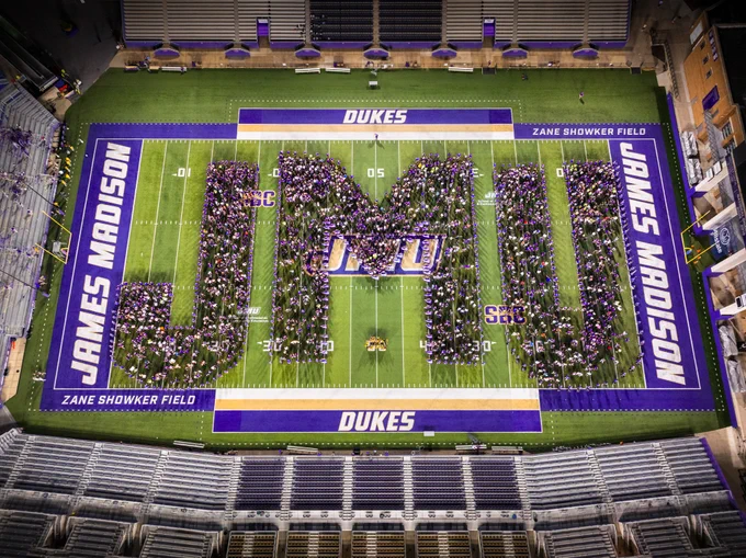 Aerial photo of "J-M-U" letters spelled out with students in formation on the field of Bridgeforth Stadium at dusk. The formation stretches from endzone to endzone.
