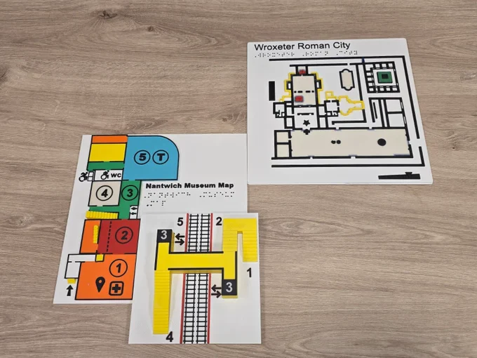 Three Direct Access MiniMaps spread out on an office table. One is for Wroxeter Roman City, another is for Nantwich Museum.  The smallest does not have a site name. All the maps are colourful, square shaped with 3D printed braille to their face.