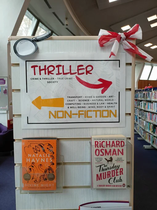Photograph of the end of a shelf. On the shelf is a poster with arrows pointing to the Thriller section on the right and the Non-fiction section on the left. Underneath the poster are two books in plastic holders. The books are Divine Might by Natalie Haynes and The Thursday Murder Club by Richard Osman. The corners of the shelf are decorated with a paper magnifying glass and a bow made of red and white striped tape. 