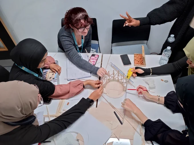 Four individuals collaborating on building a structure with wooden sticks and connectors around a circular base at a workshop table. Various crafting materials are scattered across the table, and they appear engaged in a focused group discussion.