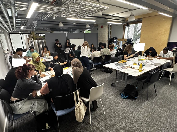 Group of individuals engaged in a workshop, sitting at tables filled with papers and craft supplies, in a room with exposed ceiling pipes and bright lighting.