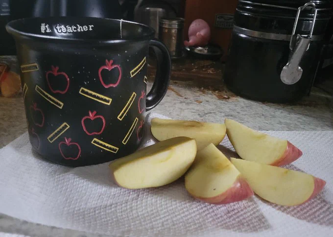Picture on kitchen counter. On left black mug with apples and pencils patterned around the exterior. On white napkins to the right is a sliced red apple.