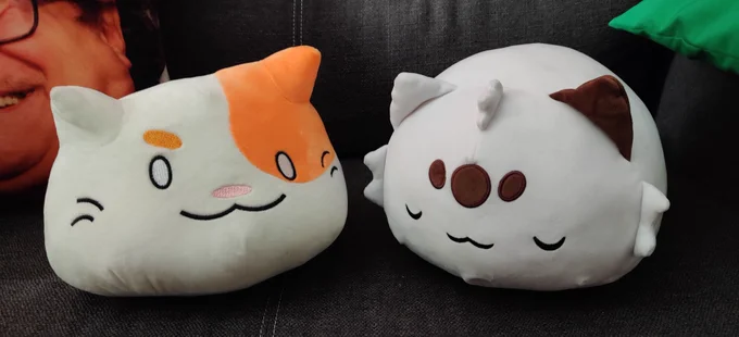 photograph of two round cat plushies; BlobCat (left) & Chipflake (right) sitting on a black couch. partially cropped off of the image is a pillow with the face of a famous actor on it for some reason