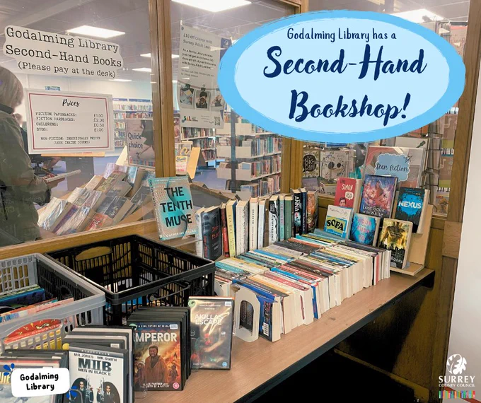 A table in the Godalming Library foyer lined with rows of books and DvDs. A caption at the top of the photo says 'Godalming Library has a second-hand bookshop!' The Godalming Library and Surrey County Council logos are in the corners of the image. 