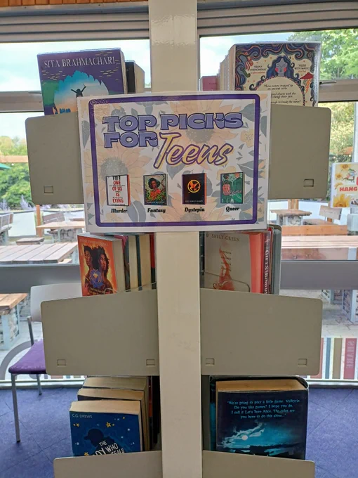 Photograph of the edge of a set of shelves with books visible on them. There is a poster promoting "Top Picks for Teens", with book cover graphics of One of Us is Lying by Karen McManus, This Poison Heart by Kalynn Bayron, The Hunger Games by Suzanne Collins, and Hearstopper by Alice Oseman. Text underneath each book cover reads "Murder", "Fantasy", "Dystopia, and "Queer" respectively. 